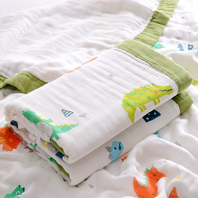  Folded 6-layer muslin cotton swaddle blanket with green trim and dinosaur print, laid on a matching muslin blanket